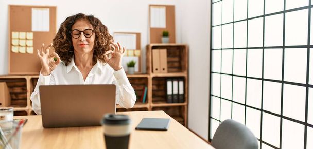 Eine hispanische Frau mittleren Alters, die im Büro arbeitet, trägt eine Brille und lächelt mit geschlossenen Augen und macht Meditationsgesten mit den Fingern. Yoga-Konzept.  - Foto, Bild