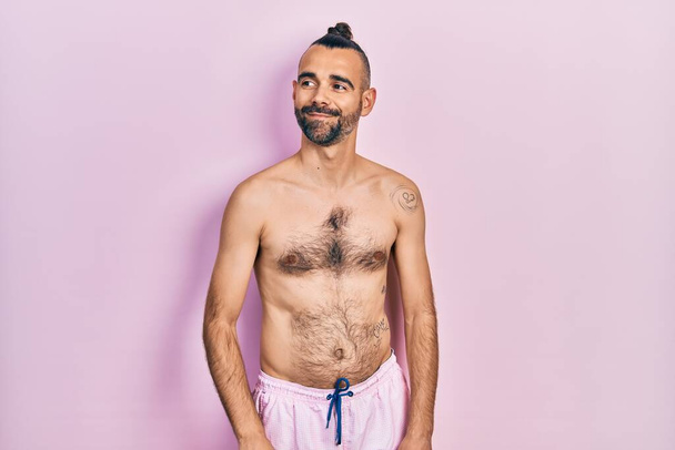 Jonge Spaanse man zonder shirt in een zwempak glimlachend naar de zijkant kijkend en wegkijkend denkend.  - Foto, afbeelding