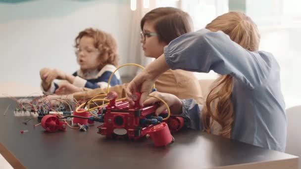 Plan moyen de trois élèves caucasiens assis ensemble au bureau pendant la leçon de technologie robotique et construisant des robots à l'aide de différents détails et fils en plastique - Séquence, vidéo