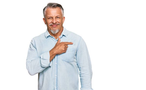 Grauhaariger Mann mittleren Alters in lässiger Kleidung, fröhlich mit einem Lächeln im Gesicht, das mit der Hand und dem Finger zur Seite zeigt, mit fröhlichem und natürlichem Ausdruck  - Foto, Bild