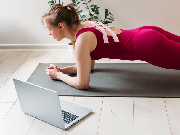 Girl training at home, doing plank. She is watching video on the internet and repeating the tasks. Indoor and outdoor workout. Sport and healthy active lifestyle concept. - Photo, Image