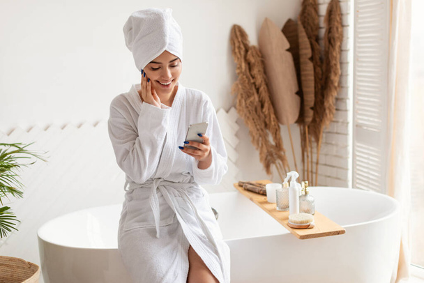Woman Using Smartphone In Bathroom Wearing Bathrobe Sitting On Bathtub - Photo, Image