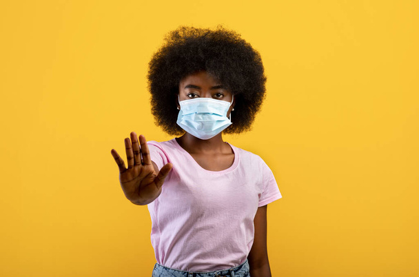 アフリカ系アメリカ人の女性は、病気に苦しみ、マスクを着用し、社会的距離を遵守し、黄色の背景に立って - 写真・画像