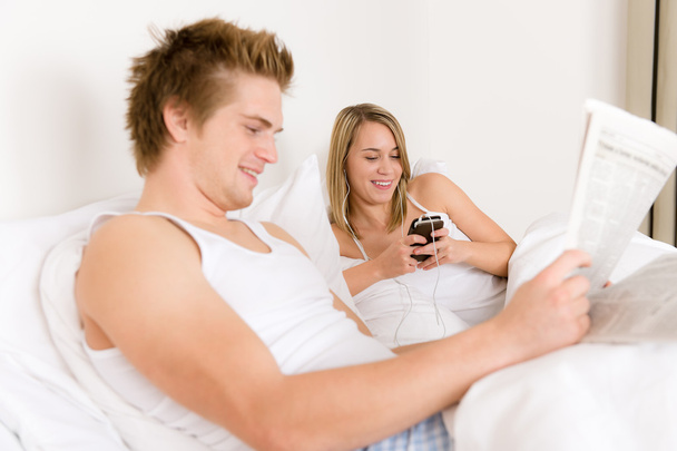 Young couple relax in white bed listen to music, read newspaper - Valokuva, kuva
