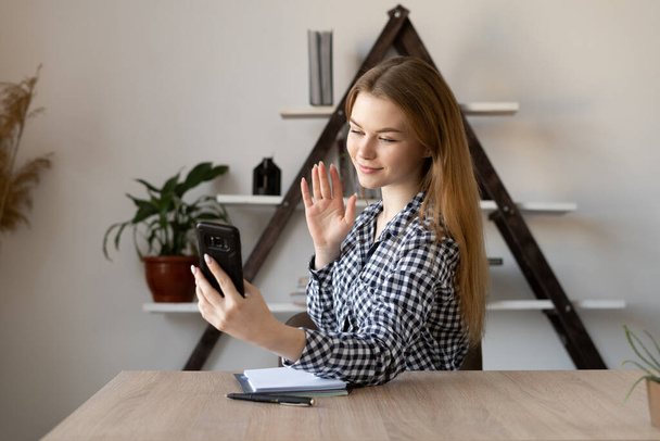 Smiling Caucasian young woman waves at smartphone camera hello and records video vlog about life. Millennial lady girl is sitting in the office and watching a video greeting. - Photo, Image
