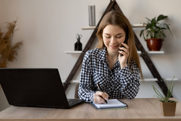 Smiling attractive young woman with a mobile phone in her hands and a laptop open on the table works remotely online from home. the call center operator accepts the clients application. - Photo, Image