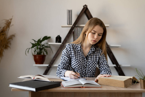 Serious focused Caucasian college girl studying at home sitting at a table with open books. European girl teacher tutor to write in a notebook teaches online remotely and prepares for a call. - Photo, image
