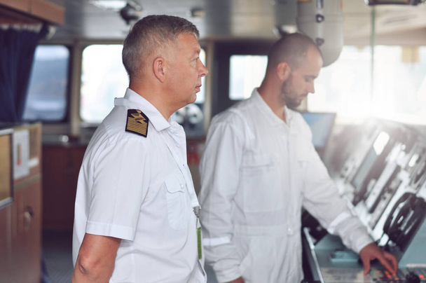 El capitán de un buque de carga con una camisa blanca y tirantes en el puente da instrucciones al navegante - Foto, imagen