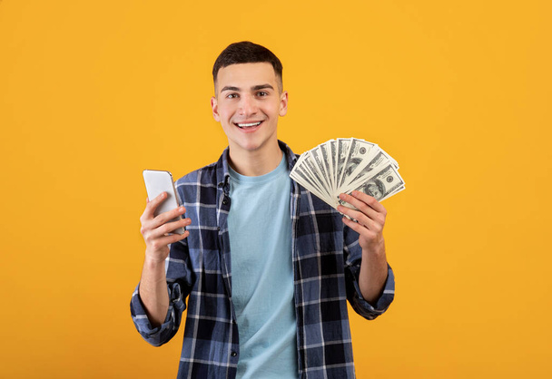 Online finances concept. Positive millennial man cellphone and American dollars on orange background - Photo, Image