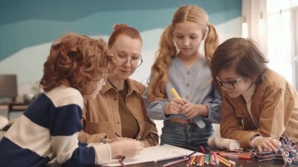 Közepes felvétel fiatal tanárnőkről, két iskolásfiúról és iskoláslányról, akiket a robotika órán gyűjtöttek össze az asztalnál, és színes filctollak segítségével rajzoltak tervrajzot papírlapra. - Felvétel, videó