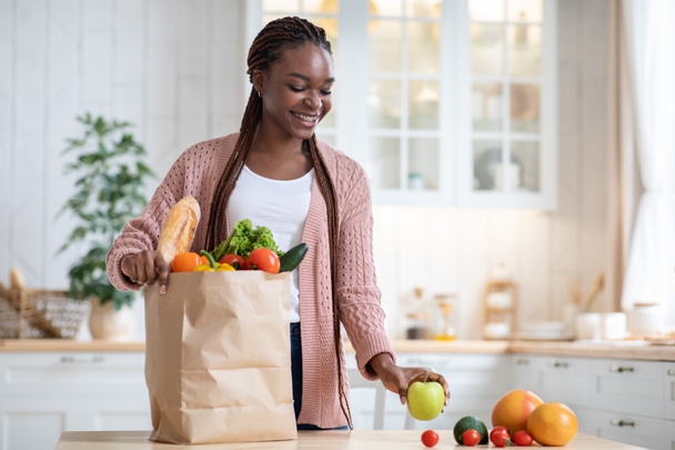 Black Lady packt nach dem Einkaufen Papiertüte mit Gemüse und Obst aus - Foto, Bild