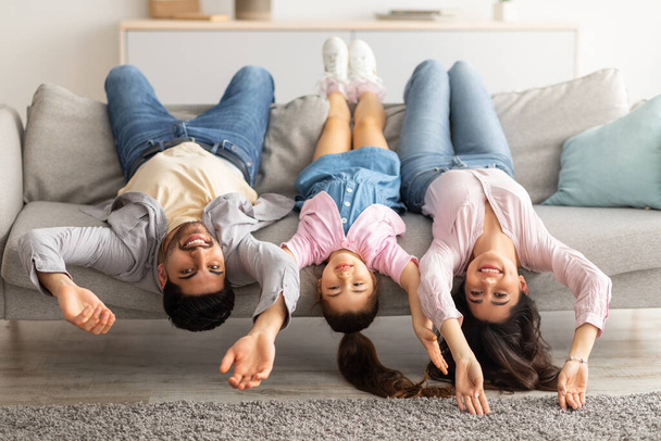 Upside down portrait of happy young family lying on sofa with heads down. Mother, father and daughter having fun - Photo, Image