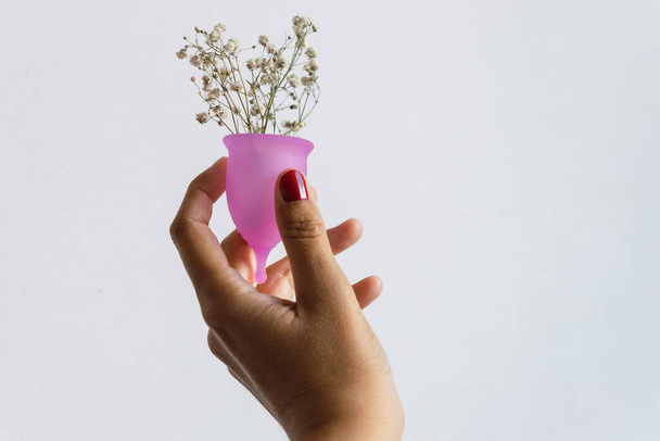 La mano femenina sostiene una copa menstrual con flores sobre fondo blanco. Higiene femenina conceptual, cuidado corporal - Foto, Imagen