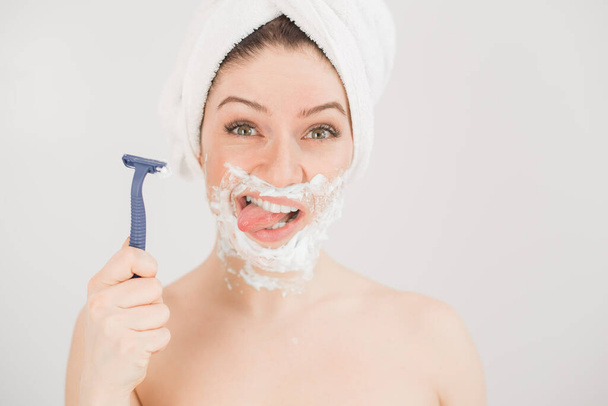 Funny portrait of a woman with shaving foam on her face holding a razor on a white background. The girl removes the mustache and beard - Φωτογραφία, εικόνα