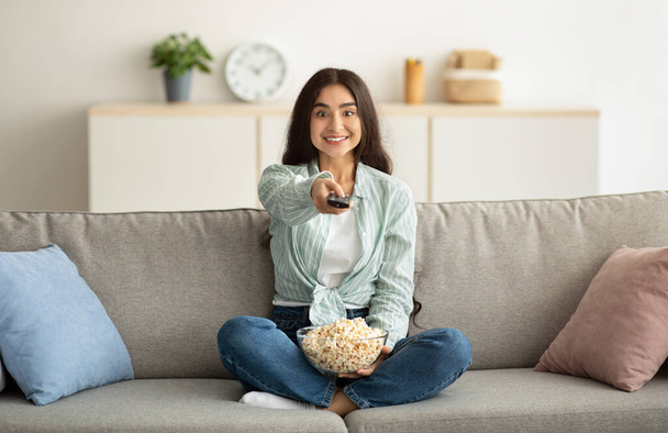 Lunghezza completa della giovane donna indiana con telecomando che guarda la TV e mangia popcorn sul divano a casa - Foto, immagini