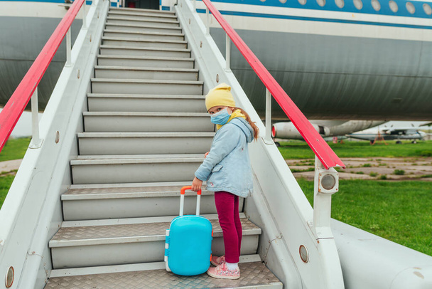 Een meisje met een beschermend gezichtsmasker met een koffer gaat naar het vliegtuig. Reizen en toerisme na de afsluiting. Nieuw normaal na Pandemic COVID-19 concept. Selectieve focus. - Foto, afbeelding