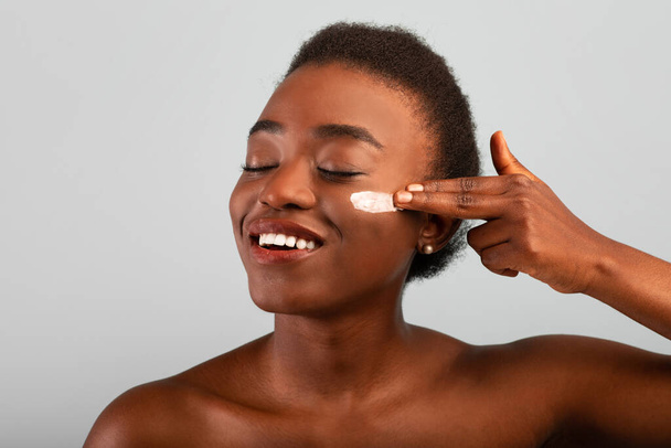 Щоденна шкіра. Африканська жінка - американка, що використовує для гідратації крем на щоці, стоїть із заплющеними очима над сірим фоном. - Фото, зображення