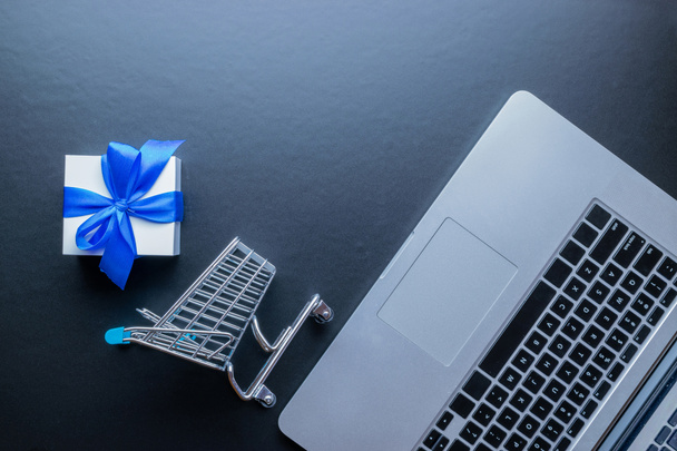 Интернет-магазины. Ноутбук компьютер, тележка и белый подарок с голубой лентой на темном фоне. Концепция электронной коммерции и клиентского опыта. Интернет покупка, интернет магазин - Фото, изображение