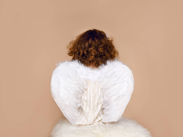 Veduta posteriore di una bambina in ali, vestita in abito seduto in studio, su sfondo beige. San Valentino. Cupidon. - Foto, immagini