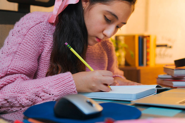 Χιλιετηρίδα έφηβος Καυκάσιος φοιτητής κάνει την εργασία στο σπίτι. Νεαρή κοπέλα κρατά σημειώσεις μπροστά από το laptop της, βιντεοκλήση από το σπίτι, σχολείο στο σπίτι - Φωτογραφία, εικόνα