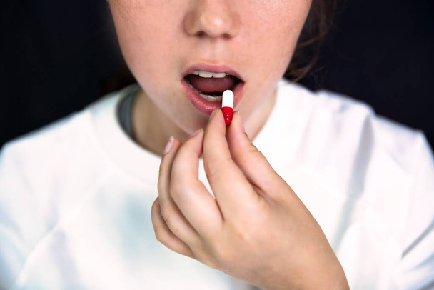 Jonge vrouw, tiener met capsule op tong op donker zwarte achtergrond, pillen slikken. geneeskunde, drugs, gezondheid concept close-up portret - Foto, afbeelding