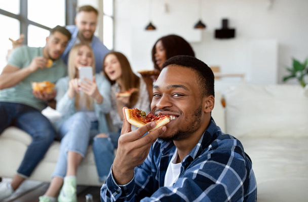 Knappe zwarte man eten plak pizza, groep van zijn multi-etnische vrienden met smartphone op de achtergrond - Foto, afbeelding