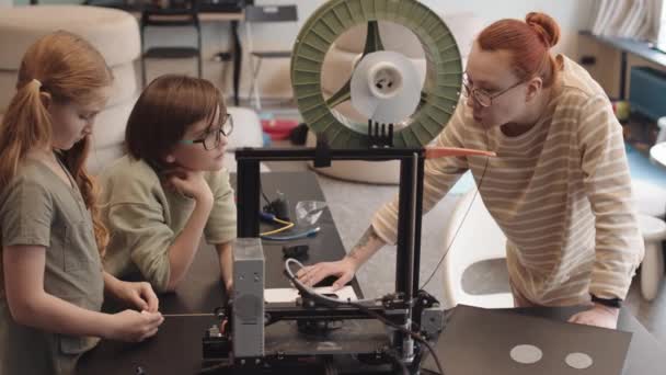 Közepes felvétel fiatal női oktatóról és két kaukázusi iskolás gyerekről, akik íróasztalnál állnak a műhelyben, beszélgetnek, miközben 3D-s nyomtatási részletek robotoknak az előtérben - Felvétel, videó