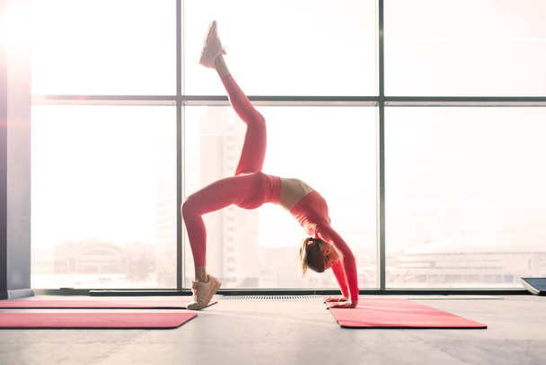 Mujer practicando yoga avanzado en estera contra una gran ventana - Foto, Imagen