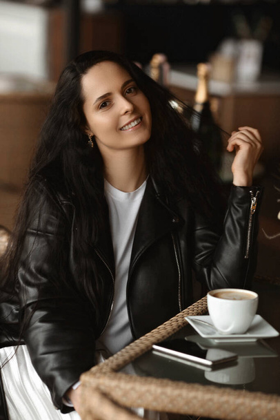 Portrait of a young female model with dark hair wearing leather jacket sitting at the table and drinking coffee in a cafe - Photo, Image