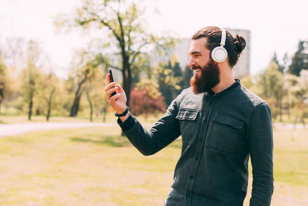 Retrato de visão lateral do homem hipster barbudo usando smartphone e usando fones de ouvido sem fio ao ar livre. - Foto, Imagem