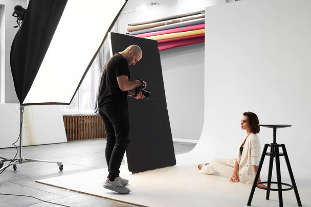 Fashion photography in a photo studio. Professional male photographer taking pictures of beautiful woman model on camera, backstage - Foto, afbeelding
