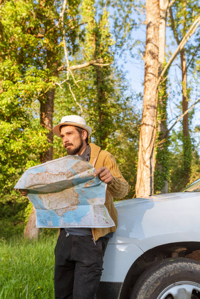 Latino man leaning on his 4wd suv car consulting on a map the route through the forest to follow. lost person with his off-road car. off-track driving. adventure concept in natural environment - Photo, Image