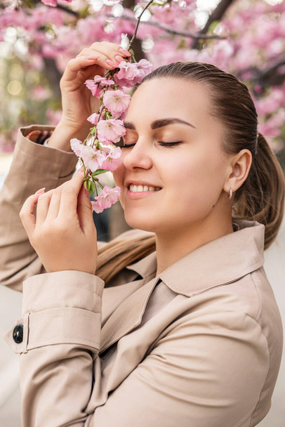 Beautiful woman near sakura trees. Pink flowers bloom in the trees on a city street. Trees bloom around. Spring time concept - Photo, Image