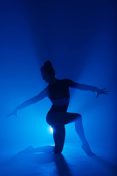 Professional ballerina dancing ballet in a smoke. Female in black bodysuit on floodlights background. - 写真・画像