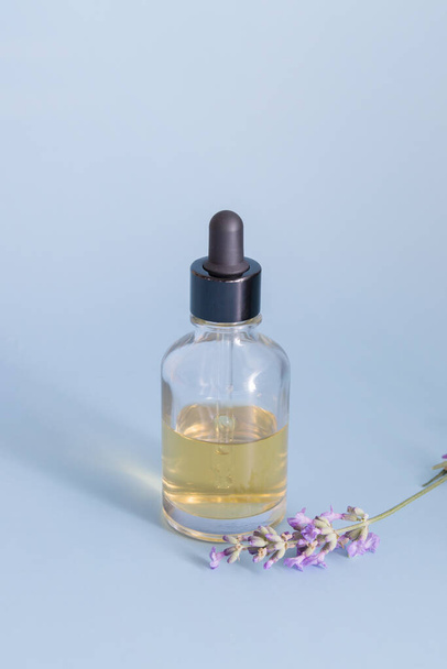 A glass bottle with a dropper filled with liquid on a blue background with lavender flowers - Foto, imagen