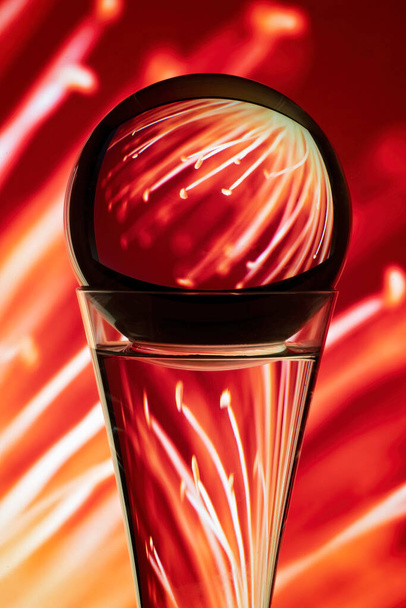 Refractie veroorzaakt door licht op reis door een ronde glazen bol bovenop een glas gevuld met water en reflecterend op zijn kleurrijke varens achtergrond. - Foto, afbeelding