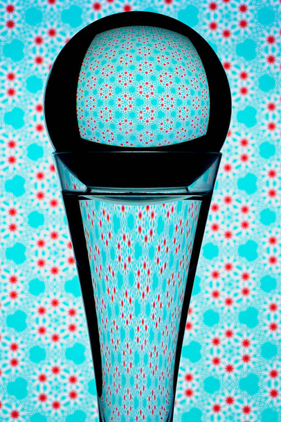 Refração causada pela luz que viaja através de um globo de vidro redondo no topo de um copo cheio de água e refletindo seu fundo padrão colorido. - Foto, Imagem