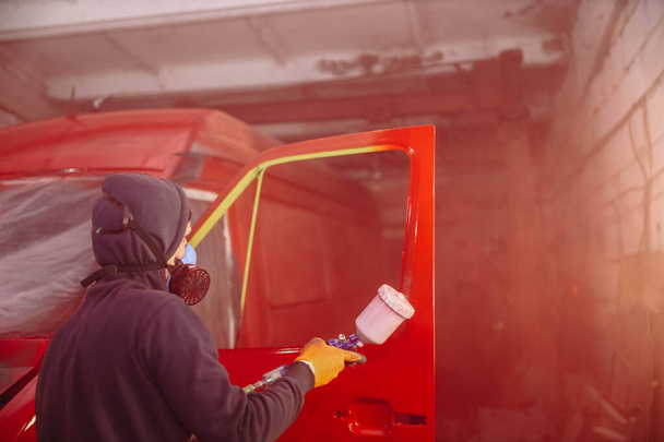man in a respirator paints a car with a spray gun in red, repair a truck body. - Foto, Imagem