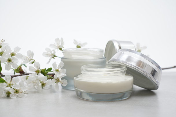 Beauty cream in glass jars on a light gray background. Decorated with white spring flowers. Unbranded skincare product. Cosmetic cream. Close up, selective focus, side view.  - Photo, Image