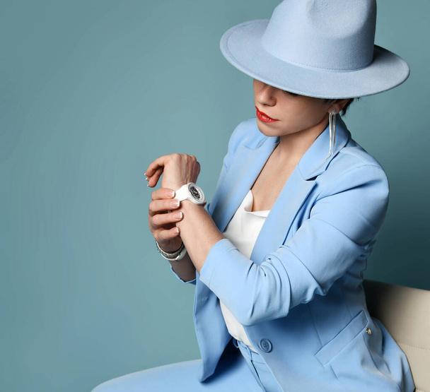 Attractive short haired brunette woman in blue business smart casual suit, hat sitting looking at watch - Valokuva, kuva