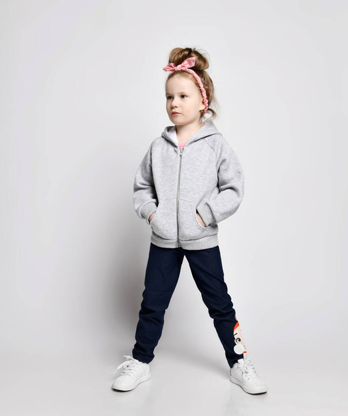 Naughty, daring, self-confident kid girl in gray hoodie with zipper standing with hands in pockets and looking aside - Fotoğraf, Görsel