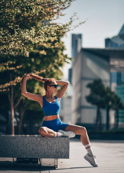 Athletic woman in sportswear, runner sits on bench form in city after workout exercises, looks aside, fixing hairstyle - Foto, imagen
