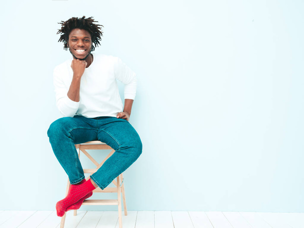 Handsome smiling hipster lambersexual model.Unshaven African man dressed in white t-shirt and jeans clothes. Fashion male with dreadlocks hairstyle sitting on chair near light blue wall in studio - Foto, Imagem
