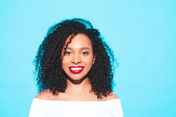 Beautiful black woman with afro curls hairstyle.Smiling model in trendy summer clothes. Sexy carefree female posing near blue wall in studio. Tanned and cheerful with red lips - Φωτογραφία, εικόνα