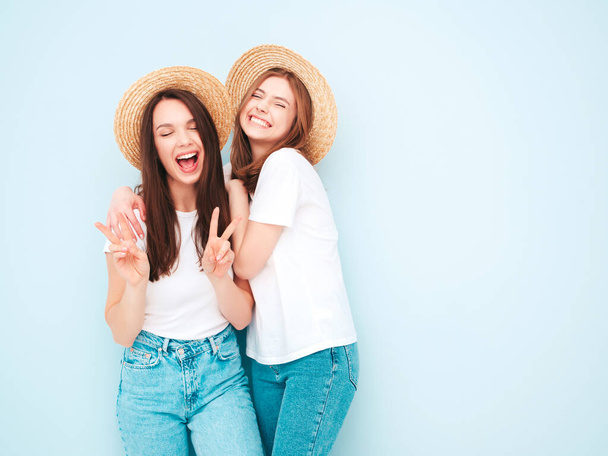 Two young beautiful smiling hipster female in trendy summer white t-shirt and jeans clothes.Sexy carefree women posing near light blue wall in studio.Positive models in hats - Photo, Image