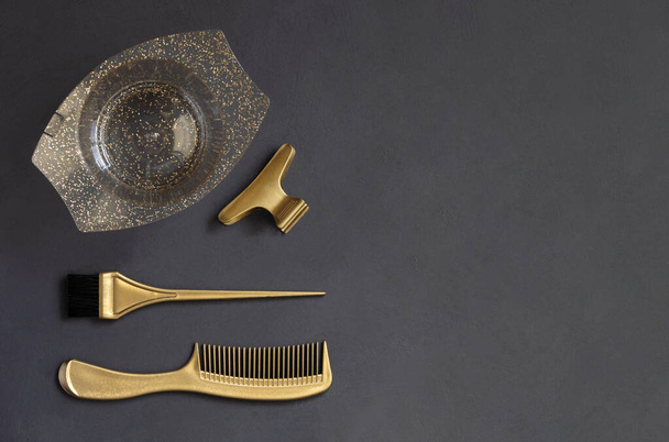 Goldene Friseurwerkzeuge zur Haarfärbung auf dunkelgrauem Hintergrund mit Kopierraum. Auf dem Tisch liegen die Accessoires des Friseursalons Kamm, Bürste, Schale und Clips, Draufsicht - Foto, Bild