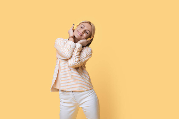 Красивое фото блондинки, слушающей музыку в наушниках, улыбающейся на желтой стене студии - Фото, изображение