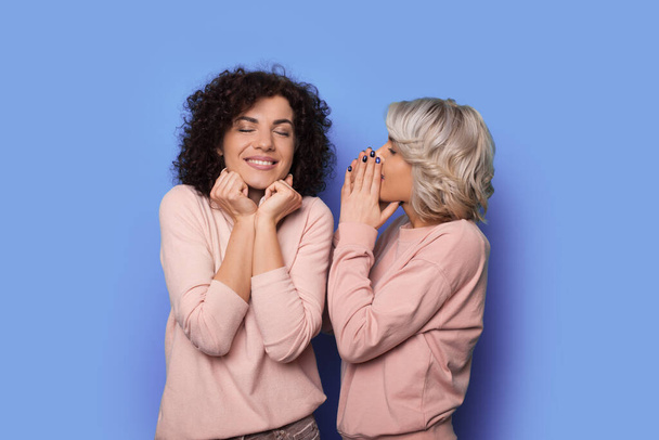 Blonde curly woman is whispering something to her brunette sister smiling on a blue studio wall - Фото, изображение