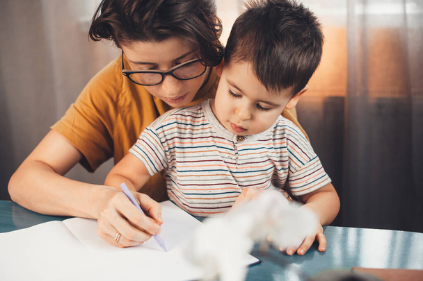 Η μελαχρινή μητέρα με τα γυαλιά βοηθάει το γιο της να μελετήσει και να γράψει γράμματα σε ένα βιβλίο. - Φωτογραφία, εικόνα