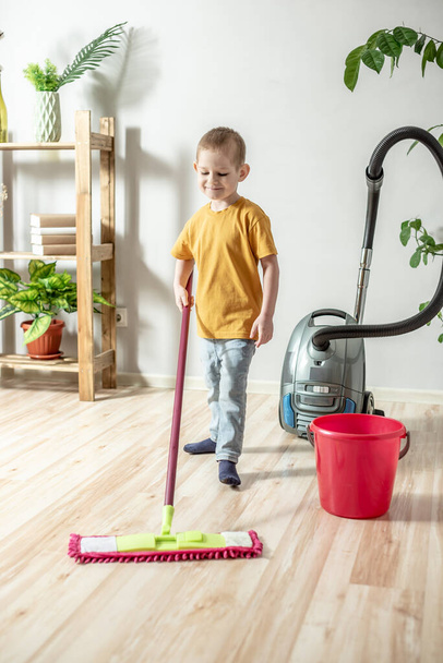 Ein kleiner Junge putzt mit einem Wischmopp den Fußboden eines Zimmers. Konzept der Selbstständigkeit, Hilfe für die Eltern, Hausarbeit des Kindes - Foto, Bild